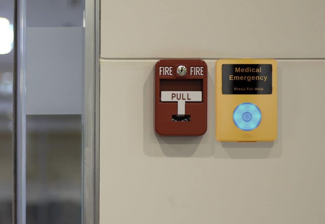 An image of an Algo 1202 call button securely mounted on a wall for emergency notification. The product is yellow with text that reads medical emergency, press for help and a big circular button that is glowing in blue. It is positioned at an accessible height next to a fire alarm.