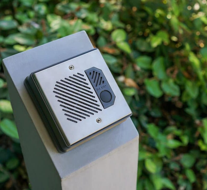 An image of an Algo 8201 door intercom system installed outside a building. The intercom unit is securely mounted on a post near the entrance. It features a speaker, microphone, and call button to initiate or accept intercom calls.